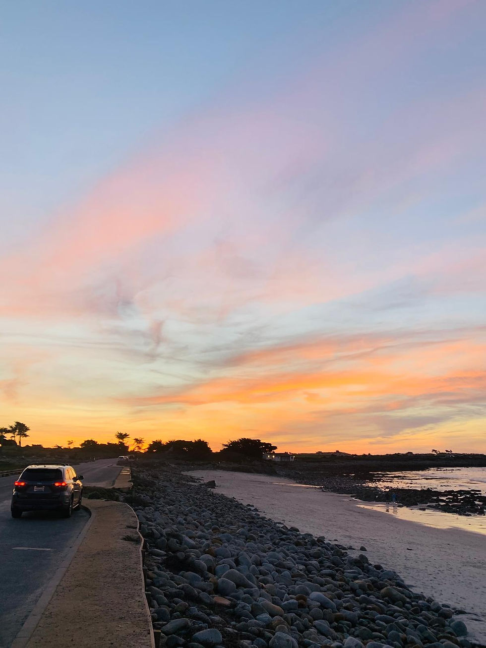 Car on coastal road at sunset with rocky beach and orange-pink sky. Calm, peaceful mood and scattered clouds in the background.