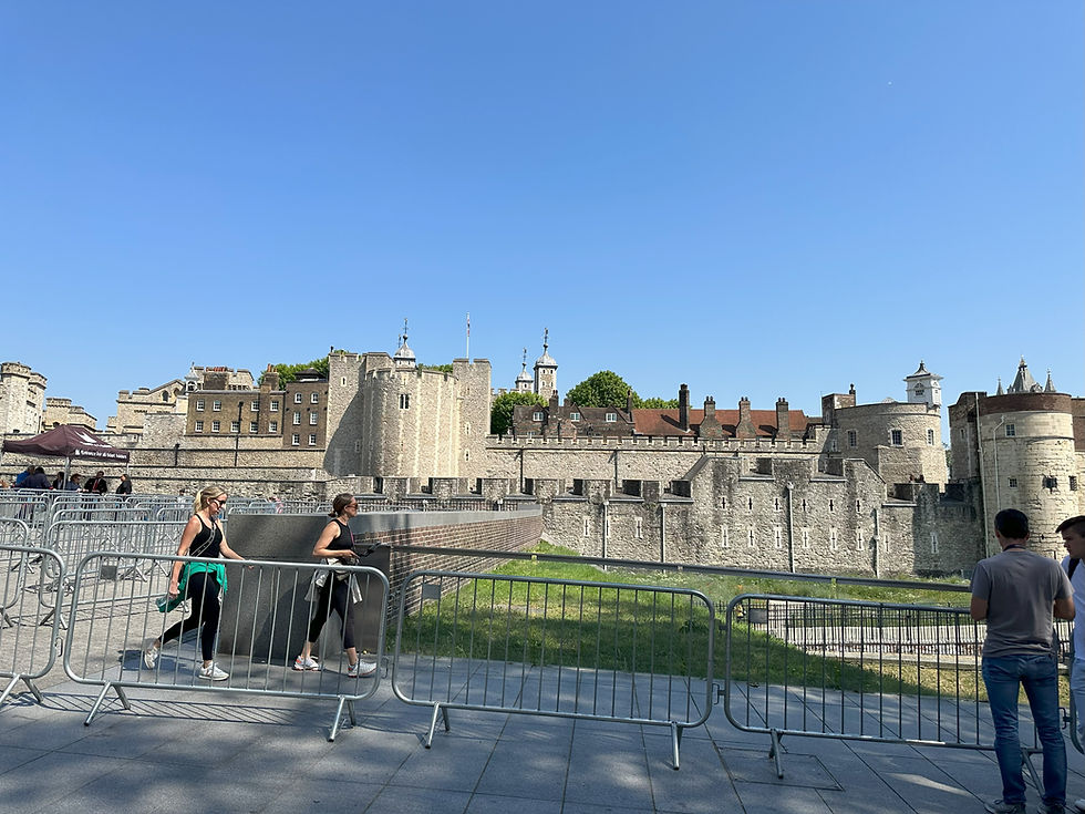 Two women walk by the Tower of London under a clear blue sky. Metal barricades line the path. People in casual attire stand nearby.