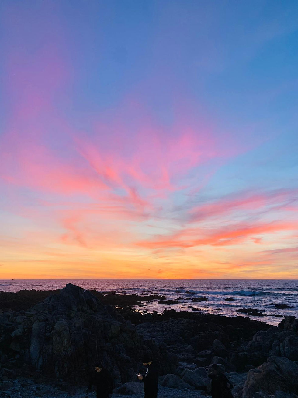 Sunset over a rocky shoreline with vibrant pink and orange sky. Two people stand in the foreground, creating a serene, contemplative mood.