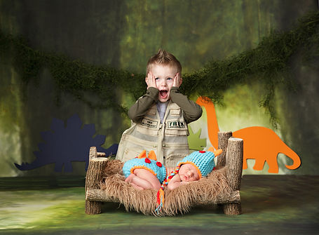 boy with his hands on his cheeks in excitement with newborn baby boy dressed as a dinosaur laying on a wood bed