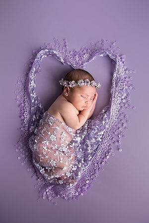 newborn baby girl on her side laying on lavender fabric with a lave fabric making a heart aroundher