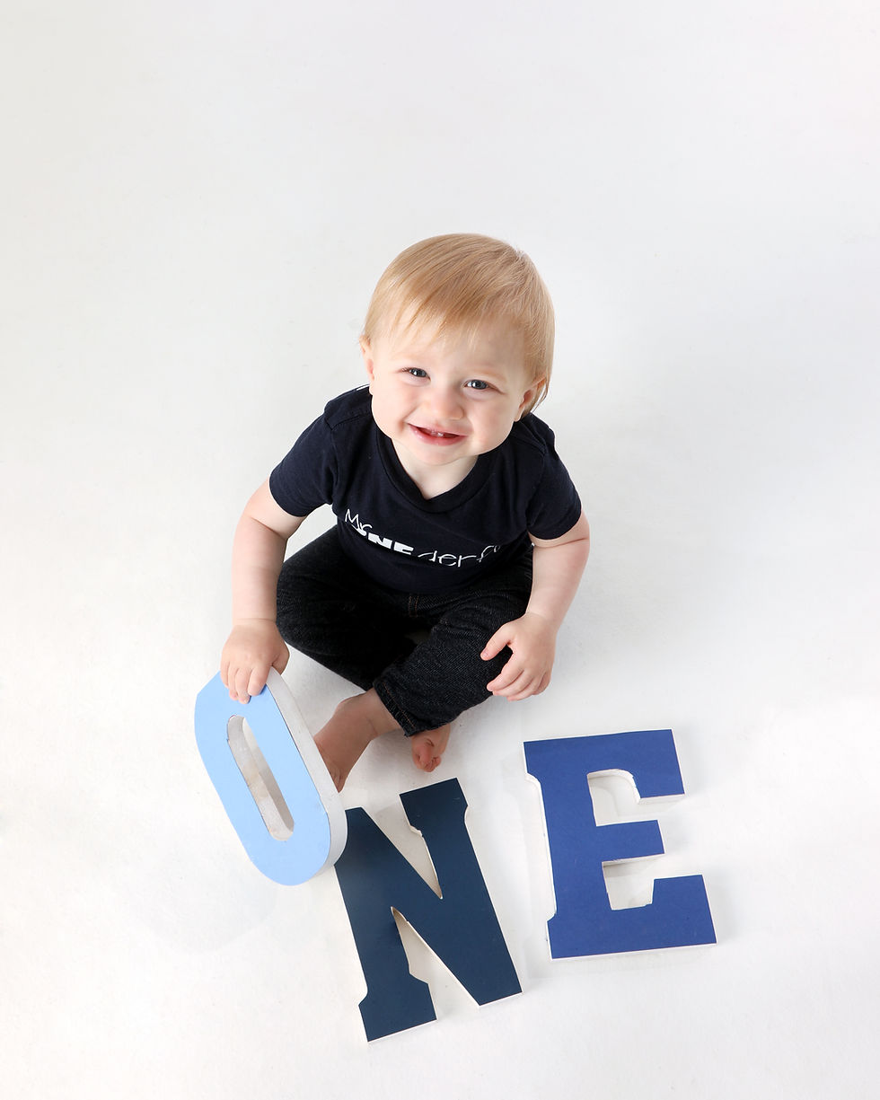 Smiling baby on white background holding a blue "O" from letters spelling "ONE" on the floor. Wears a navy shirt and black pants.