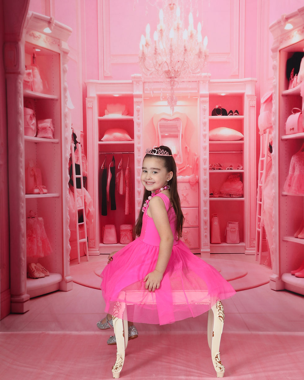 Young girl in a pink dress and tiara smiles in a pink closet setting with shelves of clothes and a chandelier above.