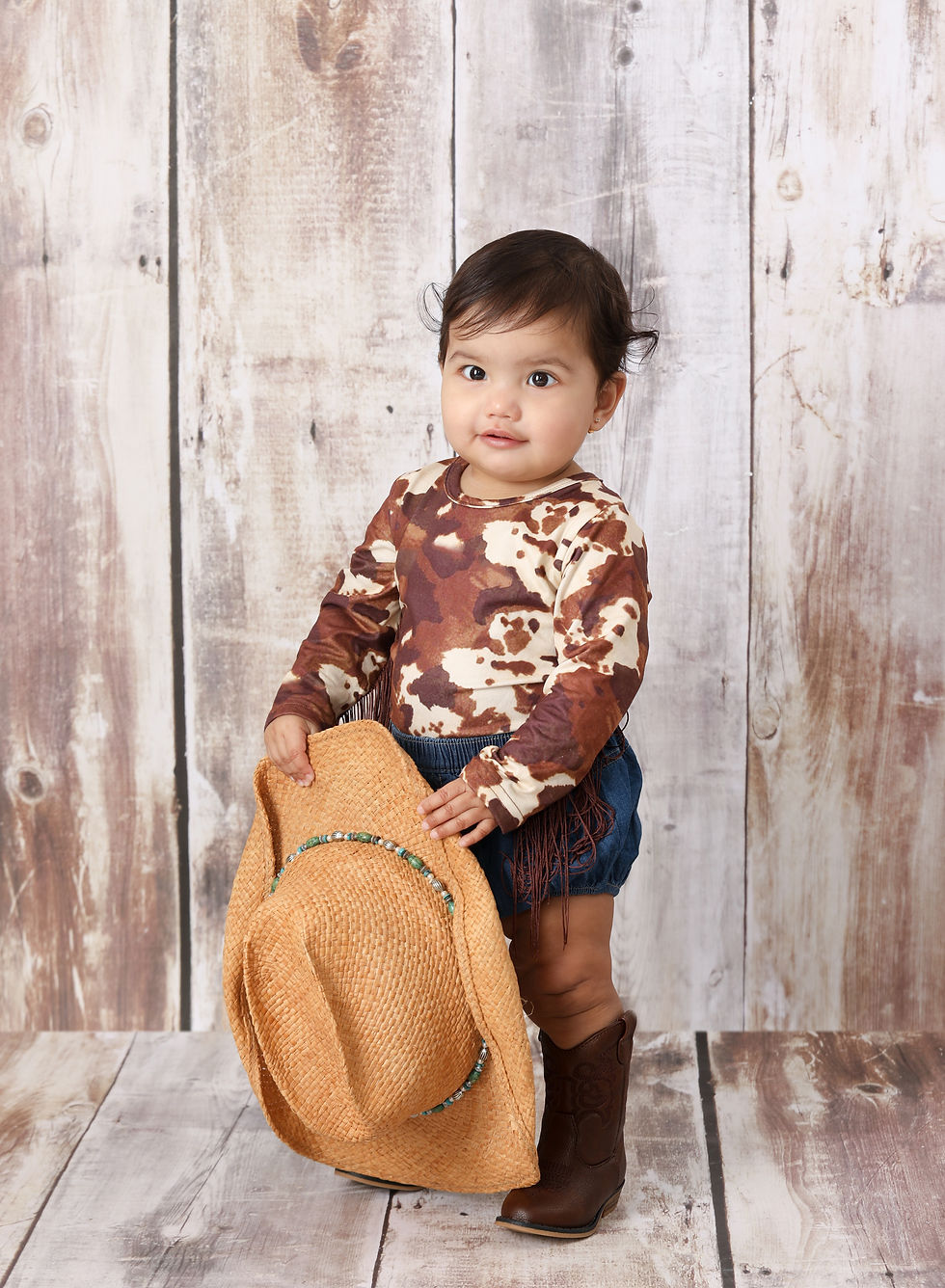 Little girl holding a cowboy hat . She is dresses in boots with a cow print shirt .