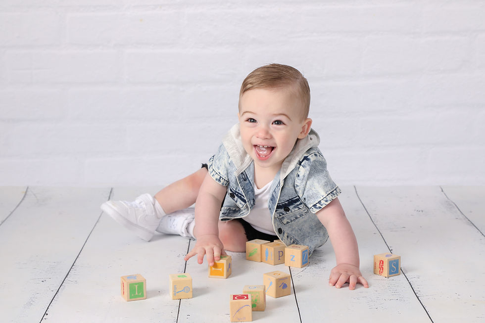 Smiling baby in denim jacket crawls on white floor, reaching for alphabet blocks in a bright studio setting.