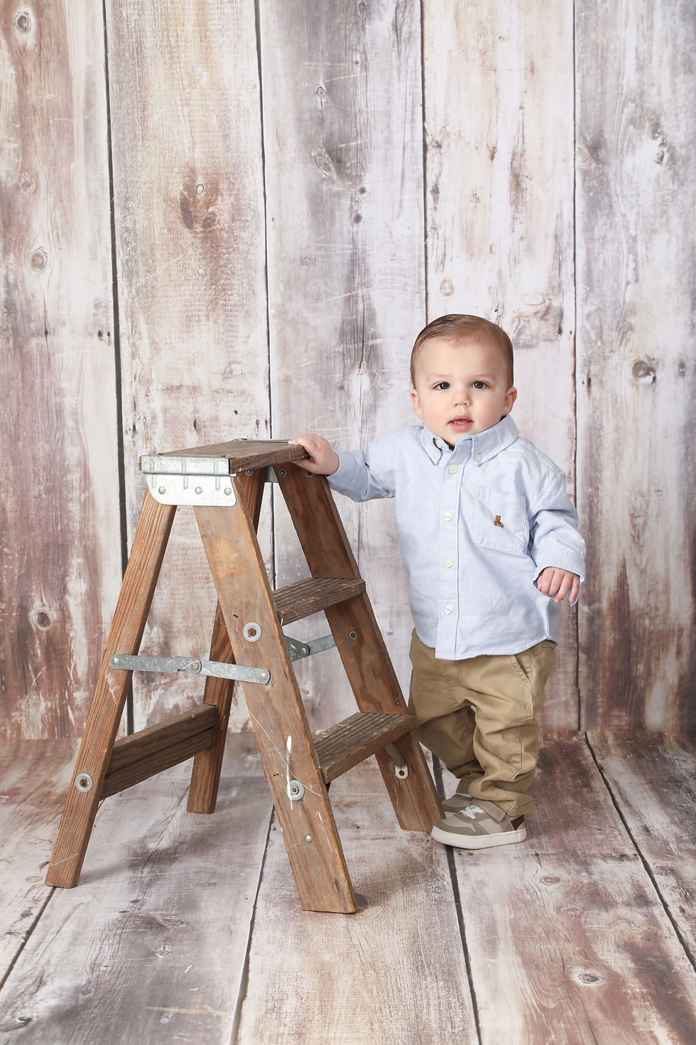 Toddler in a blue shirt and khakis stands by a wooden ladder on a rustic wood floor. Neutral tones create a calm, homey atmosphere.