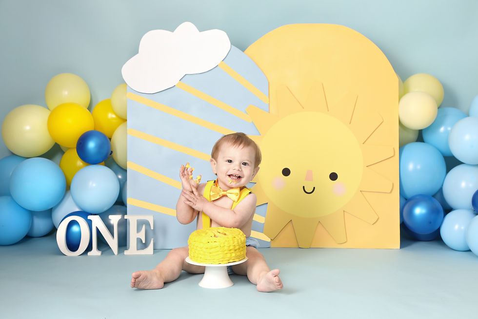 Baby with yellow cake, wearing a yellow bow tie, sits on blue floor. Smiling sun backdrop, balloons, and "ONE" letters surround them.