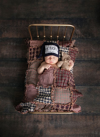 newborn baby boy laying in a brass bed with a patchwork quilt holding a teddy bear wearing a hat with his name on a wood floor