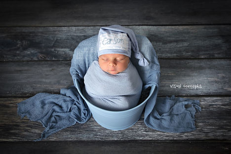 newborn baby boy in blue metal bucket swaddles in a blue wrap wearing a hat with his name on a blue wood floor