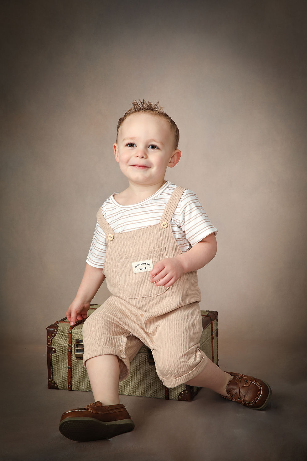 Smiling toddler in beige overalls sits on a vintage suitcase in a studio, with a small label reading Happy Every Day Child