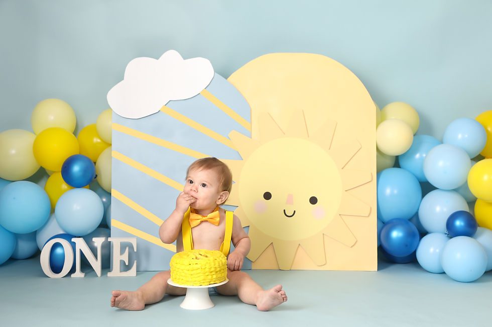 Baby with yellow suspenders and bow tie eats cake in a sunny, balloon-decorated setting. Number one displayed. Happy, playful mood.