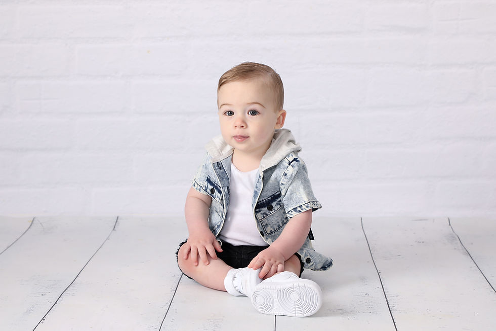 Toddler in denim jacket and white sneakers sits on a white floor, looking calmly at the camera against a plain white backdrop.