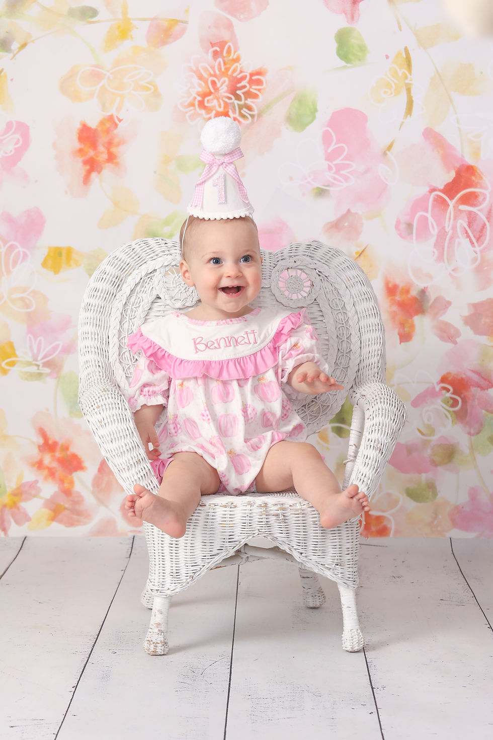 Smiling baby in pink outfit and party hat sits in a white wicker chair, with pastel floral backdrop and Bennett on her bib.