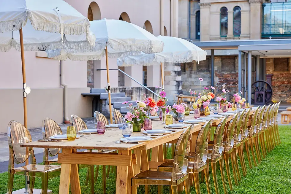 Outdoor dining setup with a long wooden table, clear chairs, colorful flowers, and white parasols on a green lawn in a historic setting.