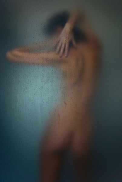Artistic, blurred photo of a nude figure pressed against textured glass, creating an abstract and ethereal effect