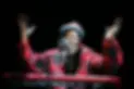 Lonnie Holley performs with arms raised at a concert in a red lumberjack coat, keyboard in front