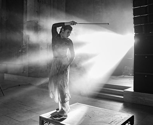Black and white photo of Lola de la Mata stood on a stage with her arm in the air pointing a violin bow to the right.