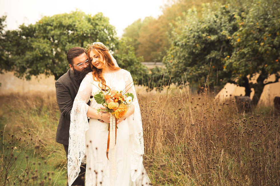 A couple embraces in a meadow, the bride holds a bouquet of orange flowers. They're smiling, surrounded by trees, with a peaceful mood.