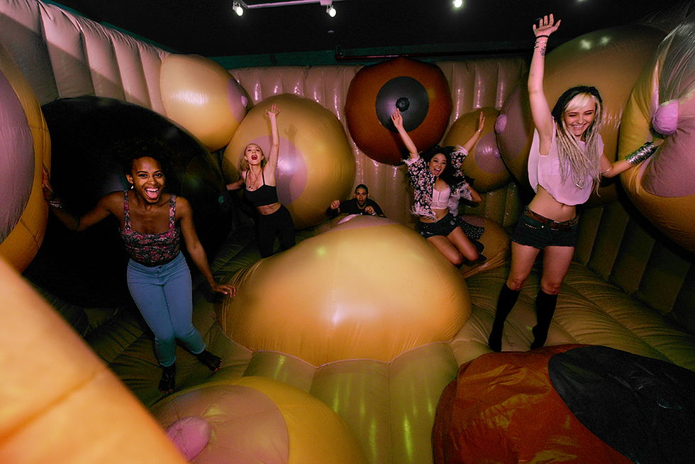 Five people joyfully bounce on a colorful, inflatable structure resembling a fun, abstract art space. The setting is lively and playful.
