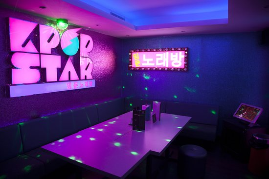 Karaoke room with purple neon "K-Pop Star" and Korean sign, large table, menus, and green laser lights. Ambiance is vibrant and playful.