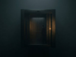A man stands in a dimly lit elevator with closed doors. A subtle orange glow highlights muted gray walls, creating a mysterious atmosphere.