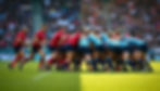 Rugby teams in red and blue uniforms scrummage on a grassy field, with a crowd in the background. Determined mood, bright daylight.