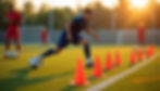 Soccer player in blue drills through orange cones on a sunlit field, as a coach observes with a tablet. Other players and goals in the background.