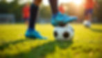 Blue soccer shoe on a ball in sunlight, grassy field, blurred players in red jerseys in the background, conveying a lively, sporty mood.