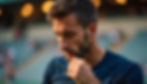 A man in a blue shirt looks thoughtful, touching his chin. Background shows blurred stadium seats. Warm light bokeh creates a calm mood.