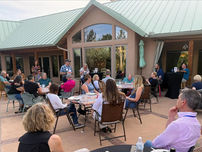 A large group of people are sitting amongst multiple patio tables listening to two people standing and talking.