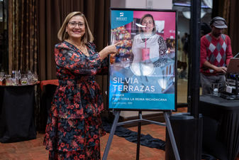 Silvia, wearing a floral dress, is smiling and pointing to a poster of her photograph as a featured client.