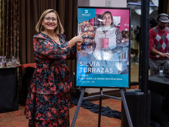 Silvia, wearing a floral dress, is smiling and pointing to a poster of her photograph as a featured client.