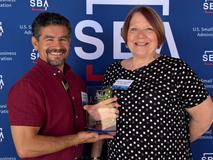 Frank and Kathleen Sanchez are smiling and standing in front of a SBA logo step and repeat banner. Frank is holding the 2026 SBA Manufacturing Small Business of the Year award.