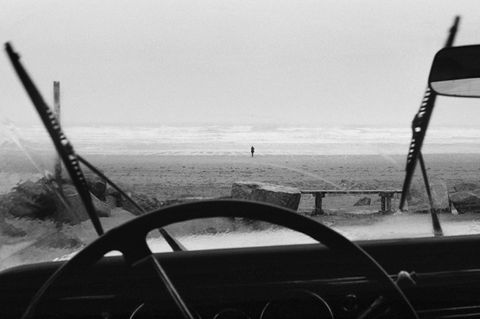 de l'intérieur d'une voiture, on voit la plage sous la pluie, des essuie glaces sont en marche, au loin une silhouette s'en va voir le rivage photo de claude pavelek
