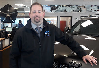 A man in front of a grey SUV wearing a black jacket with a Kootenay Auto Loans logo.