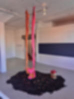 Okwae Miller's untitled piece. A swing made up of light wooden board and bright orange and hot pink fabric is suspended over a pile of dark shredded wood and wilted roses, with a paper bag filled with red roses to the right.