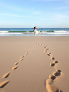 A woman in white dress twirls on a sandy beach, leaving 2 sets of footprints. Waves crash gently under a clear blue sky, creating a serene mood.