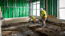 Two construction workers in full PPE levelling and pouring concrete for a new floor in a medical facility in East London.