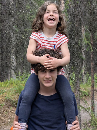 Teen carrying younger child on shoulders on forested part of Oxbow Loop Trail in Denali National Park, Alaska, surrounded by green trees
