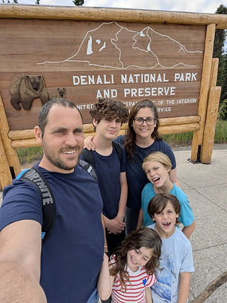 Family standing in front of the Denali National Park & Preserve wooden entrance sign, surrounded by trees and mountain scenery
