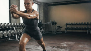 Woman in dark workout clothes lunges in a gym, focused expression. Background shows rows of dumbbells. Dim lighting, gray tones.