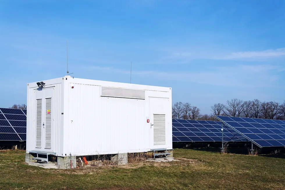 White utility building on grassy field with solar panels in background under clear blue sky. Sparse trees in distance.