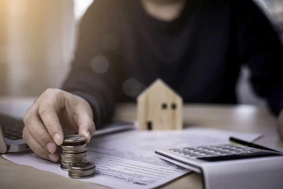 Hand stacking coins on papers beside a wooden house model, laptop, and calculator on a desk. Warm lighting creates a thoughtful mood.