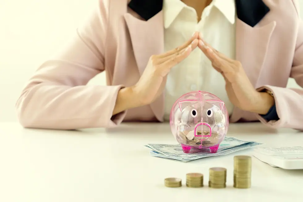 A person in a pink blazer forms a triangle with hands over a clear piggy bank on cash. Coins are stacked nearby on a white table.