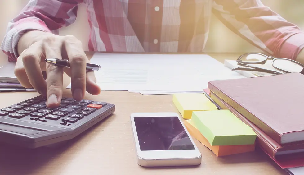 Hand using a calculator on a desk with documents, smartphone, notepads, and sticky notes. Person wears a red plaid shirt, bright setting.
