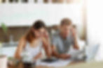 A man and woman sit at a kitchen table reviewing bills, looking concerned. There's a laptop, coffee pot, and calculator in front of them.