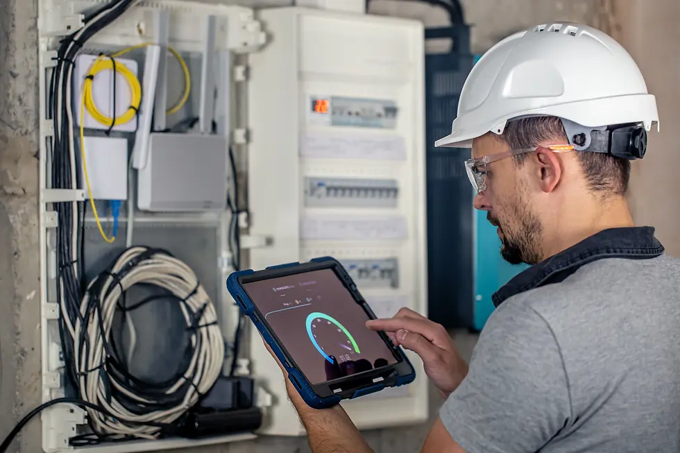 Man in a hard hat uses a tablet near an open electrical panel. Wires and cables visible. Focused expression in an industrial setting.