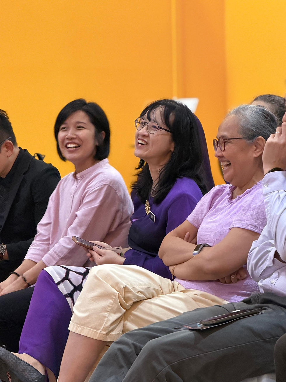 Smiling women seated indoors laugh together against a bright orange wall, wearing pastel and purple outfits in a cheerful gathering