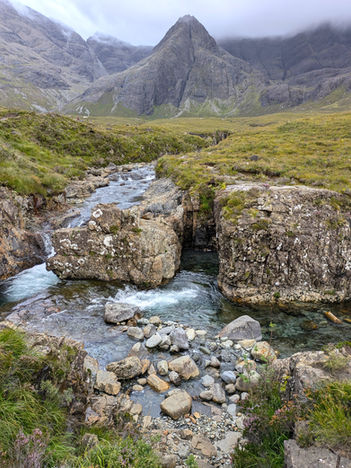 A small waterfall dropping into a clear pool of water with imposing mountains in the background making the Fairy Pools one of the best things to see and do in Skye