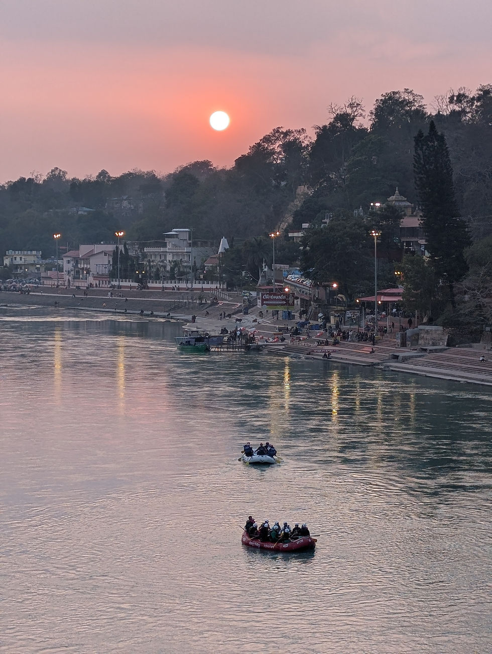 Rafts with people float on a river at sunset. The sun glows orange over a riverside town with lights, trees, and hill in the background. Places to see in rishikesh.